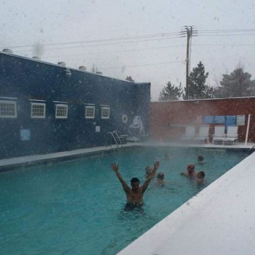 People swimming in a pool during snowfall, with snowflakes visible in the air and a cloudy sky above.