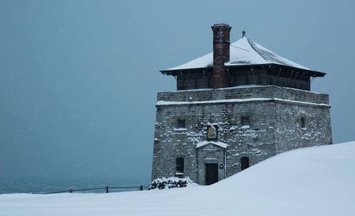 A stone building with a chimney, surrounded by snow, under a gray sky, evoking a cold winter atmosphere.