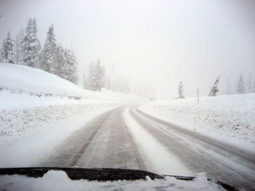 A snowy road curves through a winter landscape, surrounded by tall trees and heavy snowfall, creating a foggy atmosphere.