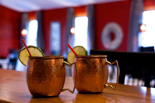 Two copper mugs with lime slices and straws sit on a wooden bar, against a red wall and softly lit background.