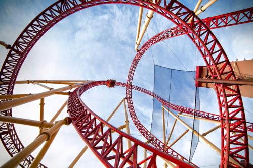 A view of a red roller coaster track twisting against a blue sky, showcasing its loops and structure.