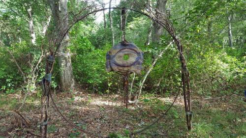 A circular wooden sculpture hangs in a wooded area, surrounded by greenery and fallen leaves.