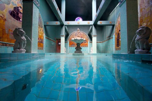 A serene indoor pool area with turquoise tiles, a central fountain, and decorative murals on the walls.