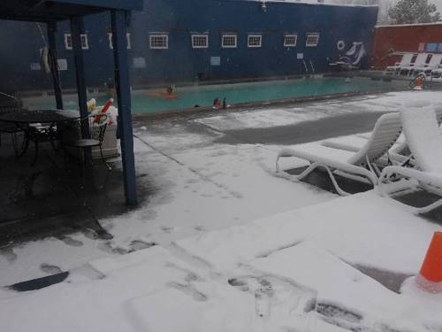 A snowy scene by an empty pool, with lounge chairs and a few people in the water, surrounded by falling snow.