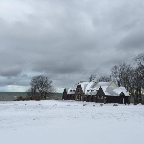 A snowy landscape featuring a dark house near a lake, with overcast skies and trees in the background.