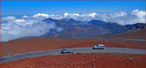 Two cars drive along a winding road above a sea of clouds, with mountains visible in the background.