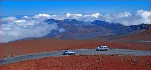 Two cars drive along a winding road above a sea of clouds, with mountains visible in the background.