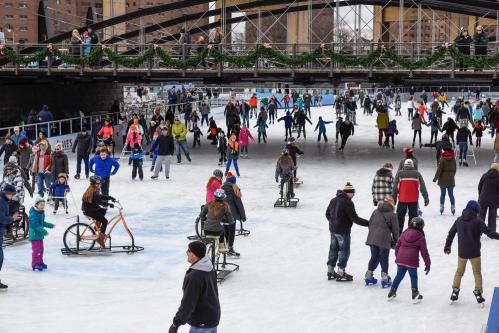 A busy ice skating rink with people skating, biking, and enjoying the winter atmosphere under a decorated bridge.