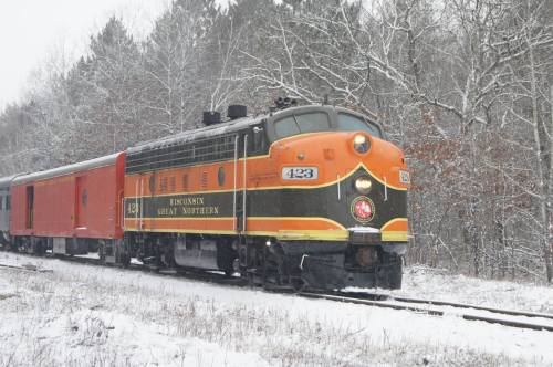 A vintage train engine in orange and black, parked on snowy tracks surrounded by trees.