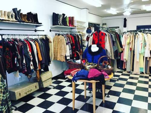 A vintage clothing store with racks of colorful garments and a table displaying accessories in a checkered floor setting.