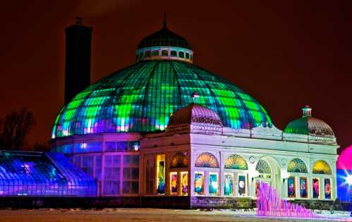 A brightly lit glass conservatory with a colorful illuminated dome against a night sky.