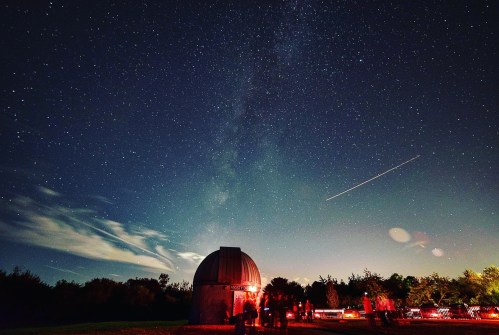 A starry night sky over an observatory, with people gathered outside and a bright streak of light in the sky.