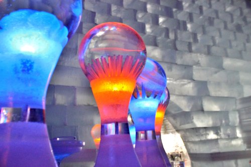 Colorful ice sculptures illuminated in blue and orange light, set against a textured ice wall.
