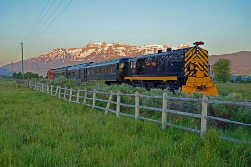 A vintage train travels through a grassy field with mountains in the background, under a colorful sunset sky.
