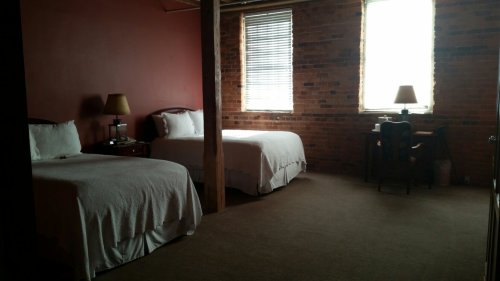 A cozy bedroom with two beds, brick walls, and a small desk by the window, featuring warm lighting and simple decor.