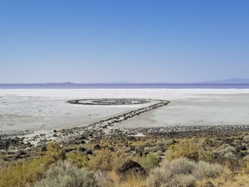 A spiral stone formation on a vast, dry salt flat under a clear blue sky. Sparse vegetation surrounds the area.