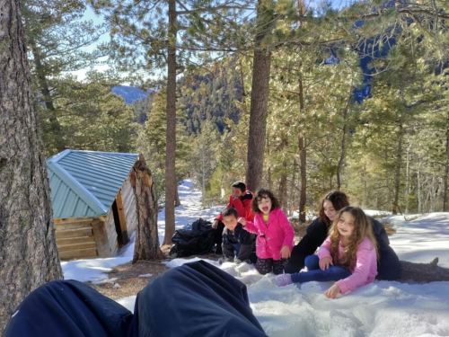 A group of children playing in the snow near a cabin, surrounded by trees and mountains on a sunny day.