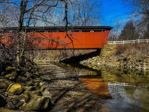 A red covered bridge spans a creek, reflecting in the water, surrounded by bare trees and rocky banks under a blue sky.
