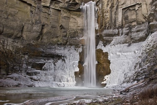 A frozen waterfall cascades down rocky cliffs into a serene, icy pool below, surrounded by rugged stone formations.