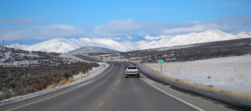 A car drives along a winding road through a snowy landscape with mountains in the background under a blue sky.