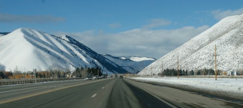 Snow-covered mountains flank a clear road under a blue sky with scattered clouds.