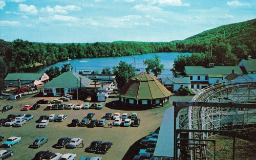 Aerial view of a lakeside amusement park with vintage cars, a carousel, and a roller coaster in the foreground.
