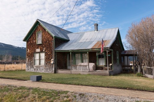 A rustic, weathered house with a metal roof, surrounded by grass and mountains under a cloudy sky. An American flag is visible.