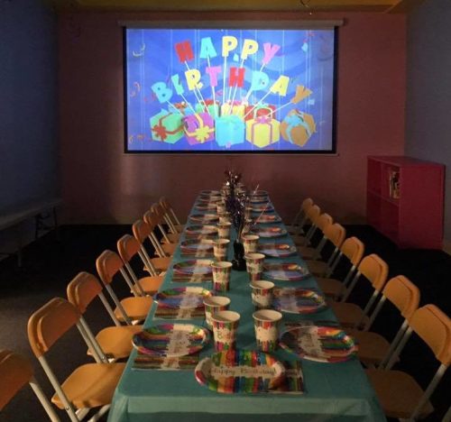 A decorated birthday party room with a long table set for guests and a "Happy Birthday" banner on the wall.