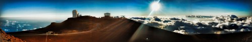 A panoramic view of a mountain peak above the clouds, with a bright sun shining in the sky.