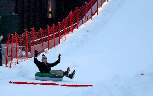 A person joyfully sliding down a snowy hill on a green inner tube, with a red safety fence in the background.