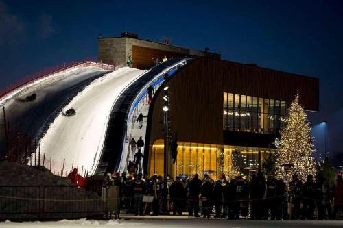 A snowy hill with people sledding at night, next to a modern building and a lit Christmas tree.