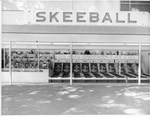 Black and white image of a skeeball arcade game setup with prizes displayed in a booth.