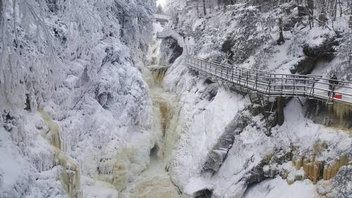 A snowy landscape featuring a waterfall cascading through a rocky gorge, with wooden walkways above.