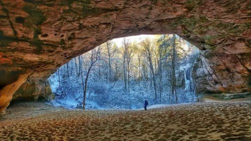 A person stands in a sandy cave entrance, overlooking a snowy forest and waterfall in the background.