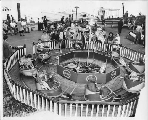 A vintage carousel with children riding in small boats, surrounded by spectators at a fair or amusement park.