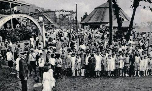 A large crowd of people, mostly children in vintage clothing, gathered at an amusement park with a roller coaster in the background.