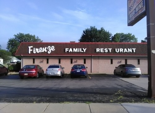 Sign for "Firenze Family Rest Urant" on a building, with several parked cars in front. Trees in the background.