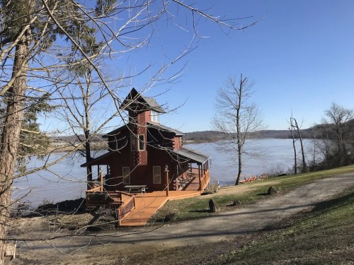 A red house by a river, surrounded by trees, with a clear blue sky and a peaceful landscape.
