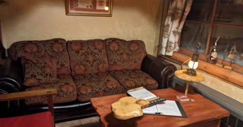 Cozy living room with a patterned couch, wooden table, lamp, and documents on a rustic surface. Warm, inviting atmosphere.