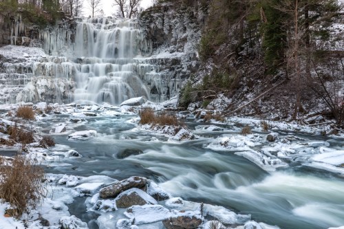 A frozen waterfall cascades down rocky cliffs, surrounded by icy water and sparse vegetation in a winter landscape.