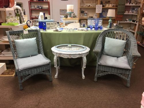 Two wicker chairs with blue cushions and a glass-top table in a store setting, surrounded by decorative items.