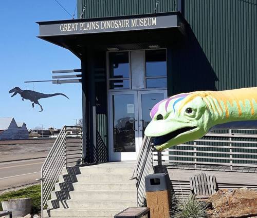 Colorful dinosaur sculpture in front of the Great Plains Dinosaur Museum, with a dinosaur silhouette in the background.