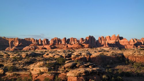 A panoramic view of rugged red rock formations under a clear blue sky in a desert landscape.