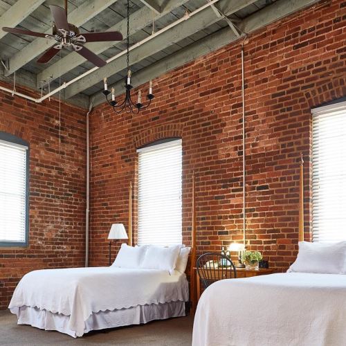 Cozy bedroom with exposed brick walls, two beds, a ceiling fan, and large windows letting in natural light.