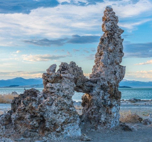Tufa formations rise from the shore of a lake, with mountains and a blue sky in the background.