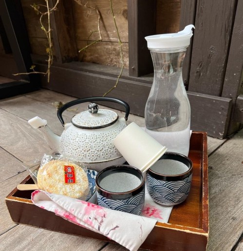 A wooden tray with a teapot, two cups, a water pitcher, and a sweet treat, set against a rustic background.