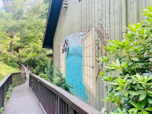 A wooden path leads to a building with a mural of open shutters and a serene blue scene surrounded by greenery.