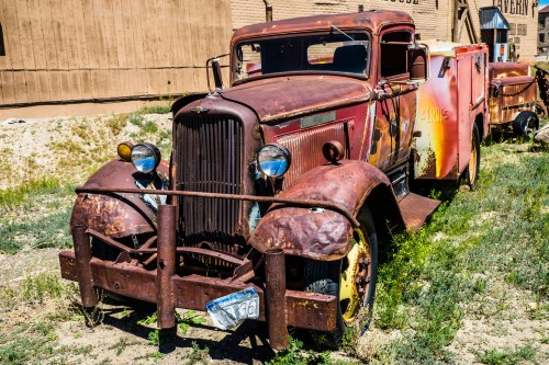 An old, rusty truck with a weathered body parked on grass, surrounded by a rustic backdrop.