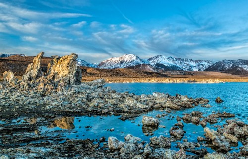 Rocky shoreline with unique formations, reflecting mountains and a blue sky over a calm lake.