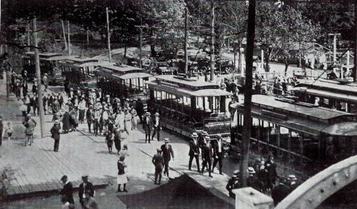 A historic black-and-white photo of a busy street with trams, pedestrians, and early automobiles.